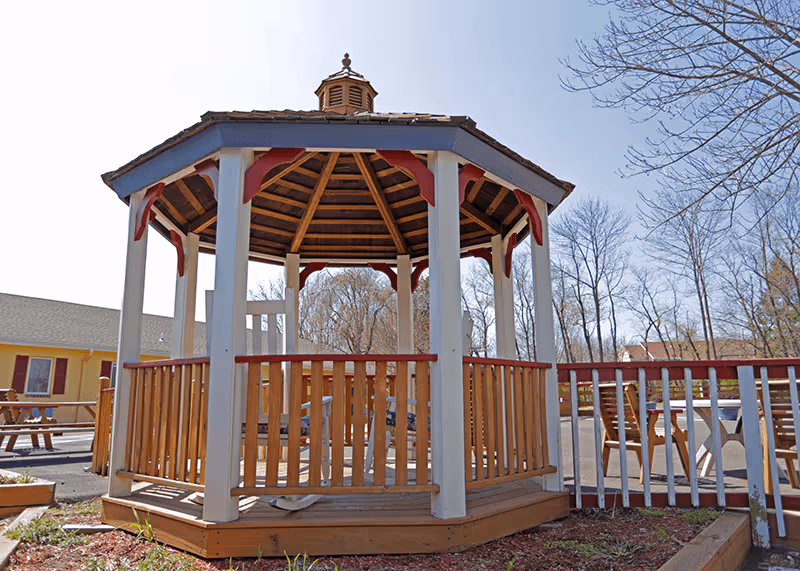 A wooden gazebo with a hexagonal roof and railings, situated outdoors on a sunny day. Surrounding the gazebo are some wooden benches and tables, with leafless trees and a building with yellow walls and red shutters in the background.