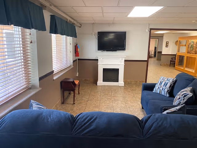 A cozy living room area with two blue sofas adorned with patterned pillows facing a white electric fireplace mounted on a beige and white wall. Above the fireplace is a flat-screen TV. The room has tiled flooring, two windows with blinds and dark green valances, and a small wooden side table near the windows. An open doorway leads to a hallway with additional seating and wooden furniture.