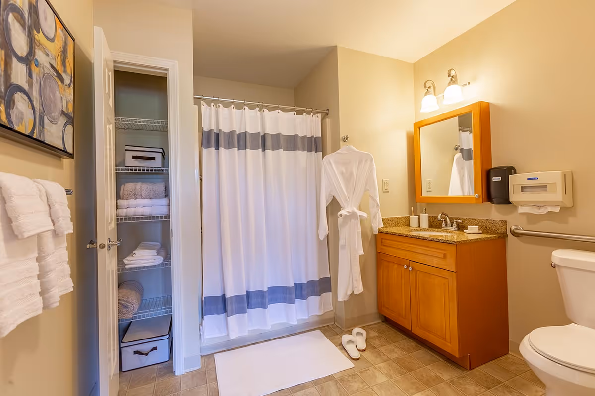 A clean and well-lit bathroom featuring a shower with a white curtain with gray stripes, a wooden vanity with a granite countertop and a mirror above it, a white bathrobe hanging on the wall, white slippers on the floor, a toilet, and an open linen closet stocked with towels and storage bins. There is also a towel rack with white towels and a piece of abstract wall art.