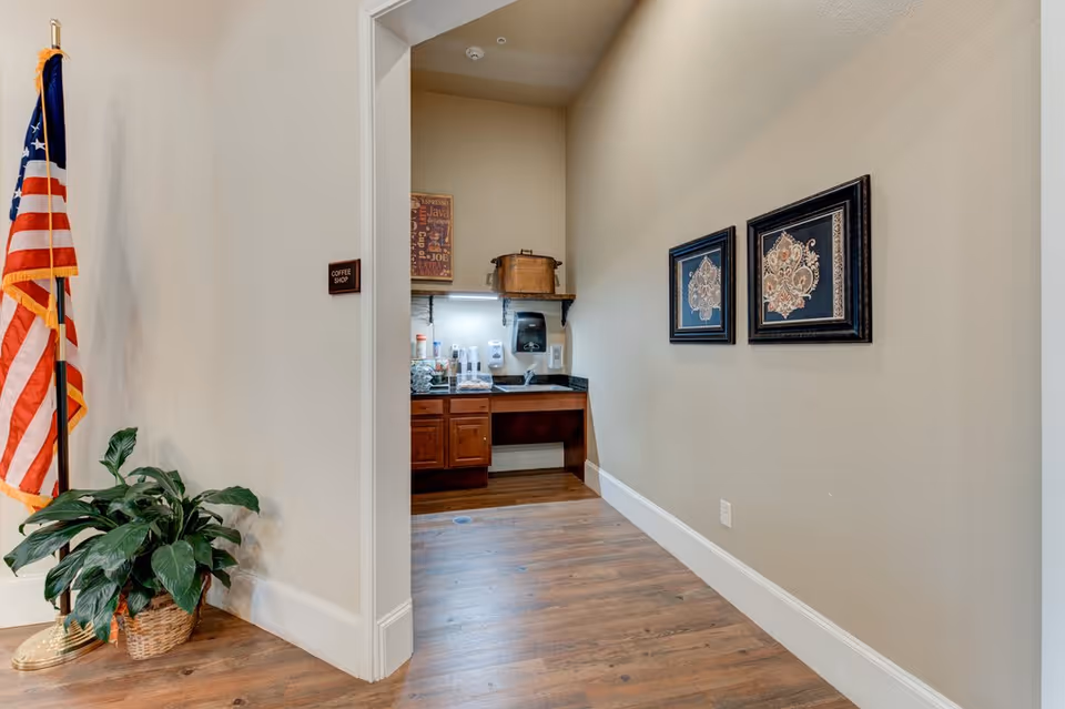 Interior view of a hallway leading to a small coffee shop area with a countertop, sink, and coffee supplies. The hallway has two framed decorative pictures on the right wall, a potted plant on the left near an American flag, and a sign on the wall indicating 'Coffee Shop'.