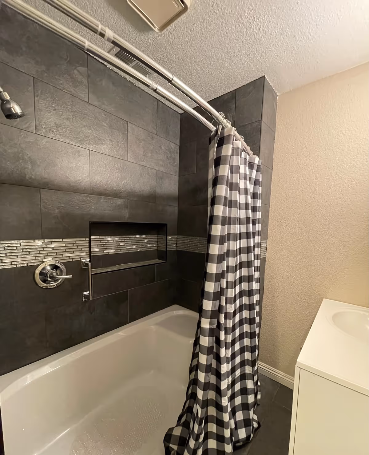 Bathroom with a white bathtub, dark gray tiled walls with a decorative horizontal strip, a silver showerhead, and a black and white checkered shower curtain. A white sink vanity is partially visible on the right side.