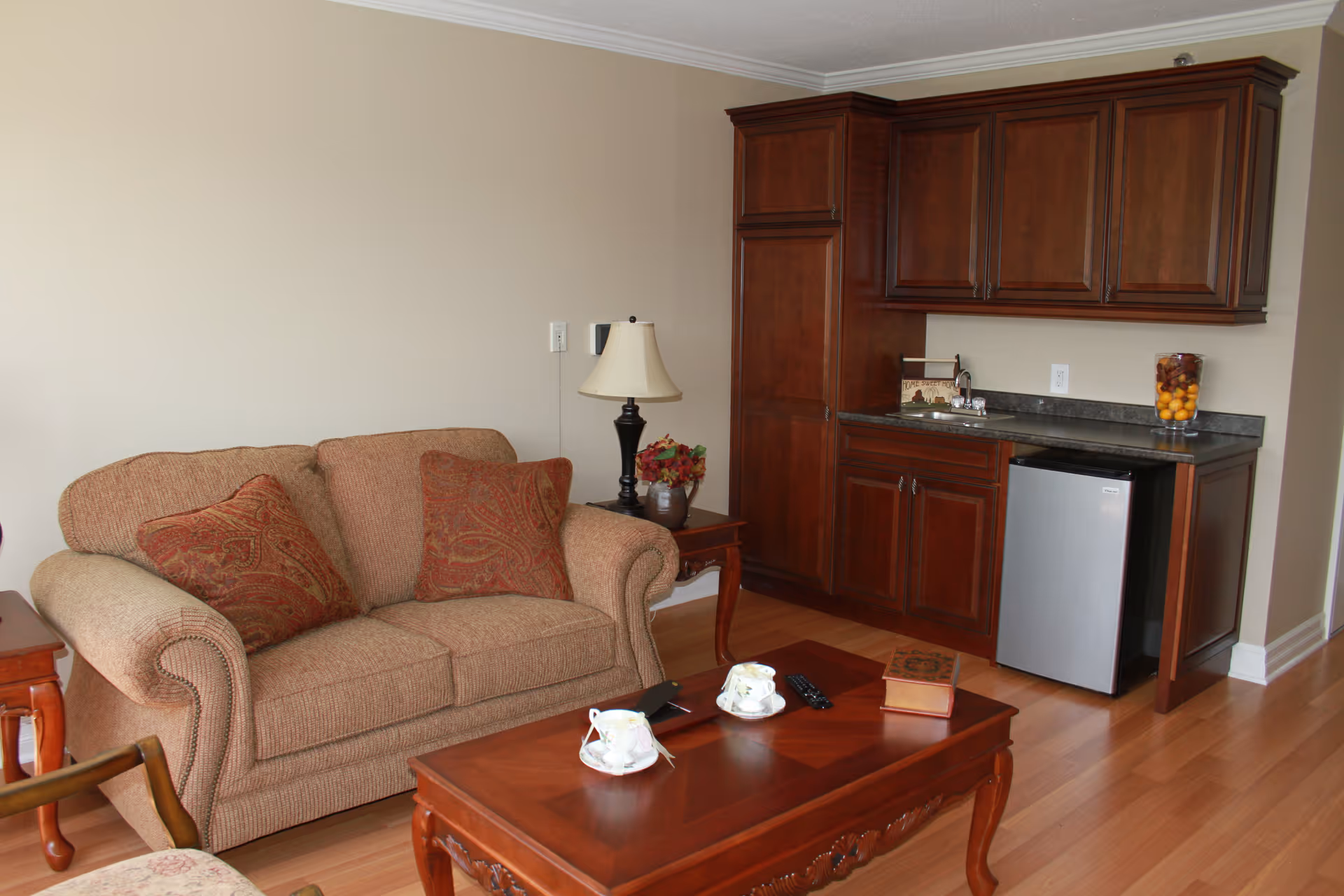 A cozy living room area with a beige loveseat adorned with two patterned cushions, a wooden coffee table with two teacups, a remote control, and a book on it. To the right, there is a wooden cabinet with a small refrigerator underneath, a countertop with a decorative jar of fruit, and a table lamp on a side table next to the loveseat.