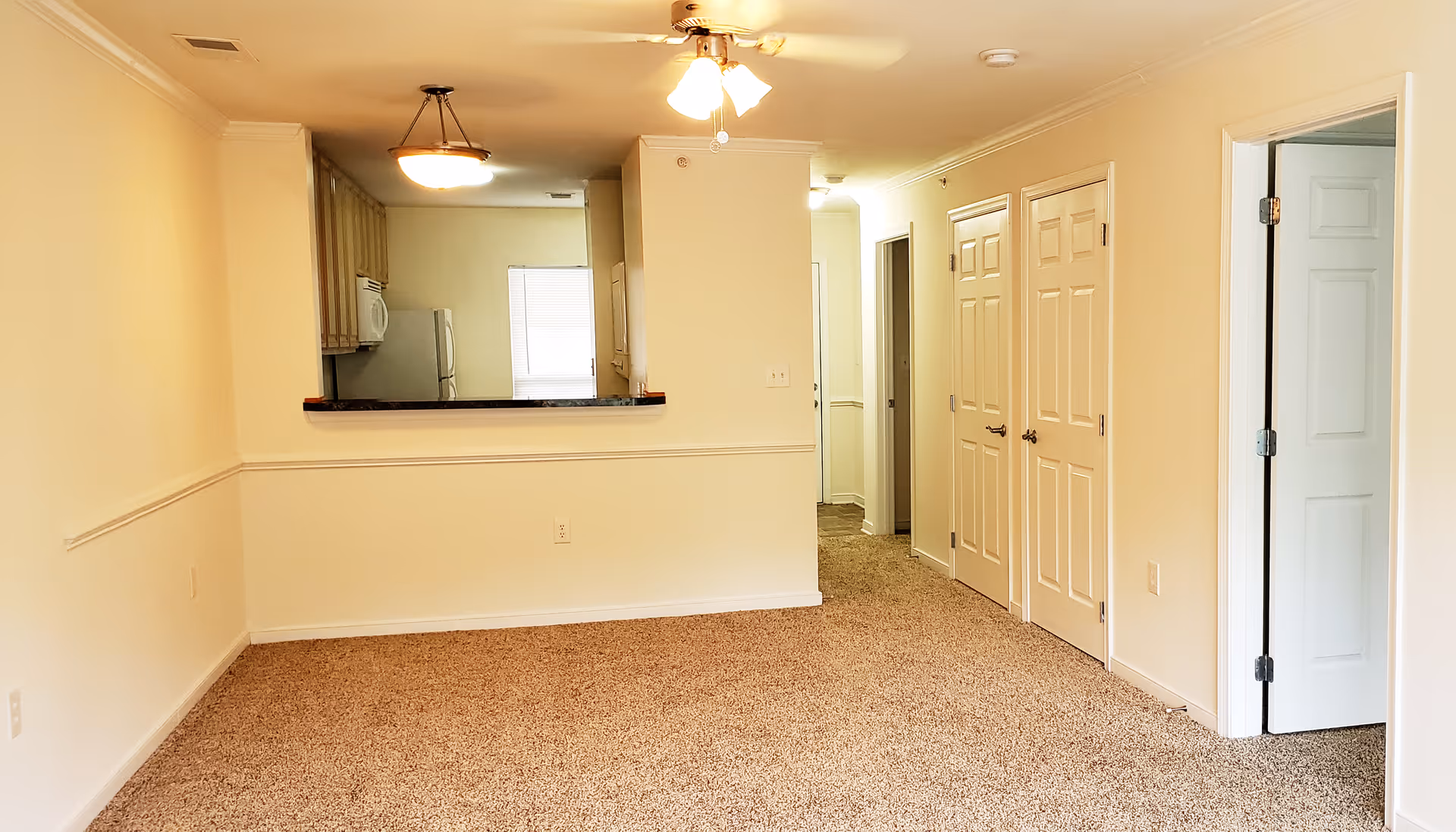 Empty room with beige carpet and cream-colored walls featuring a ceiling fan with lights, a pass-through window to a kitchen with visible refrigerator and microwave, and several white doors along a hallway.