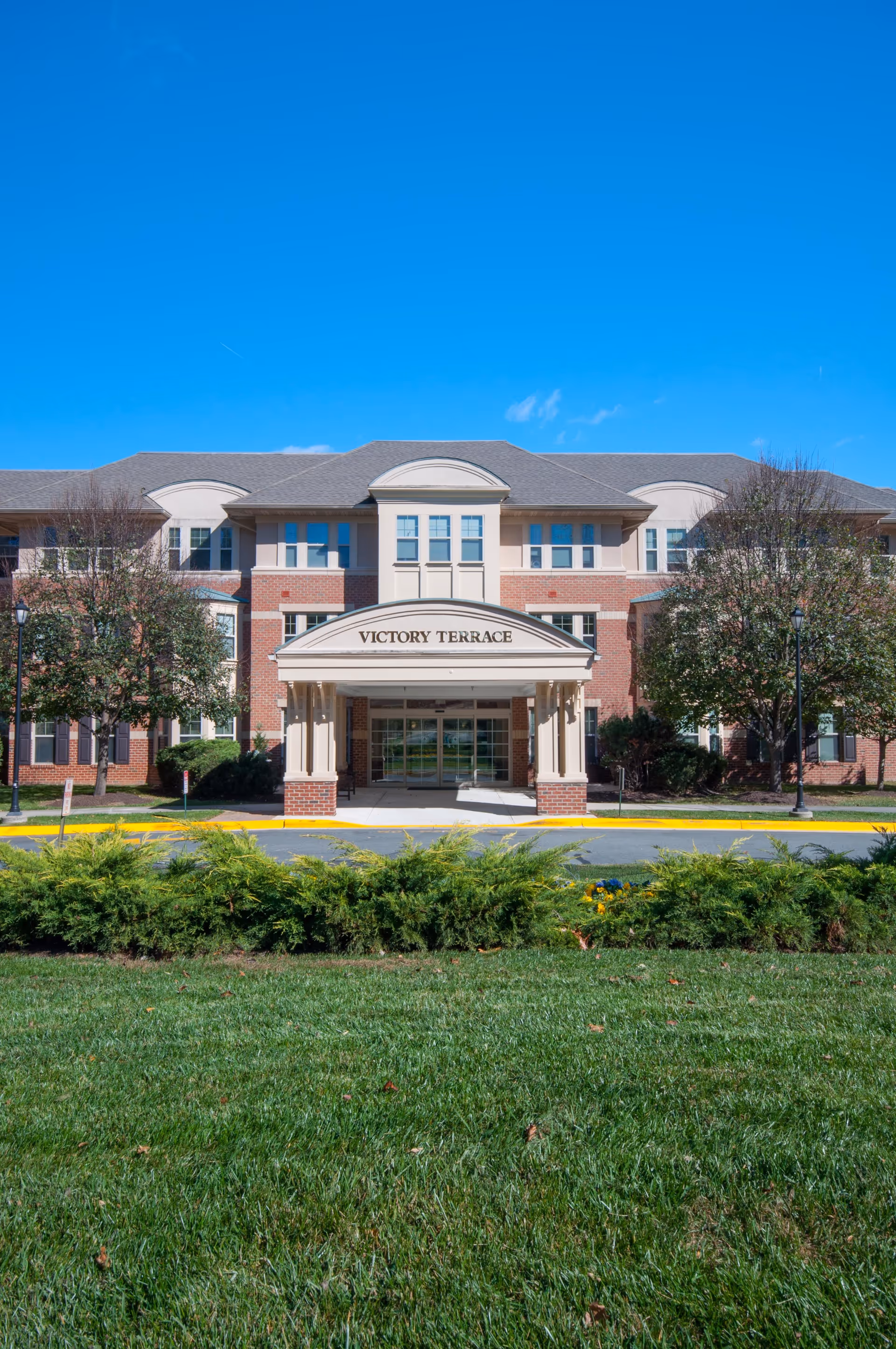 Front exterior view of Victory Terrace, a three-story senior living facility with a brick and beige facade, a covered entrance, and well-maintained landscaping including green grass, bushes, and trees under a clear blue sky.