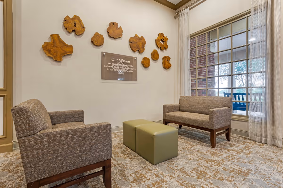 A cozy seating area in a senior living facility with two upholstered chairs and a small green ottoman in the center. The wall features decorative wooden art pieces and a framed mission statement. A large window with sheer curtains lets in natural light and shows an outdoor porch with a swing.