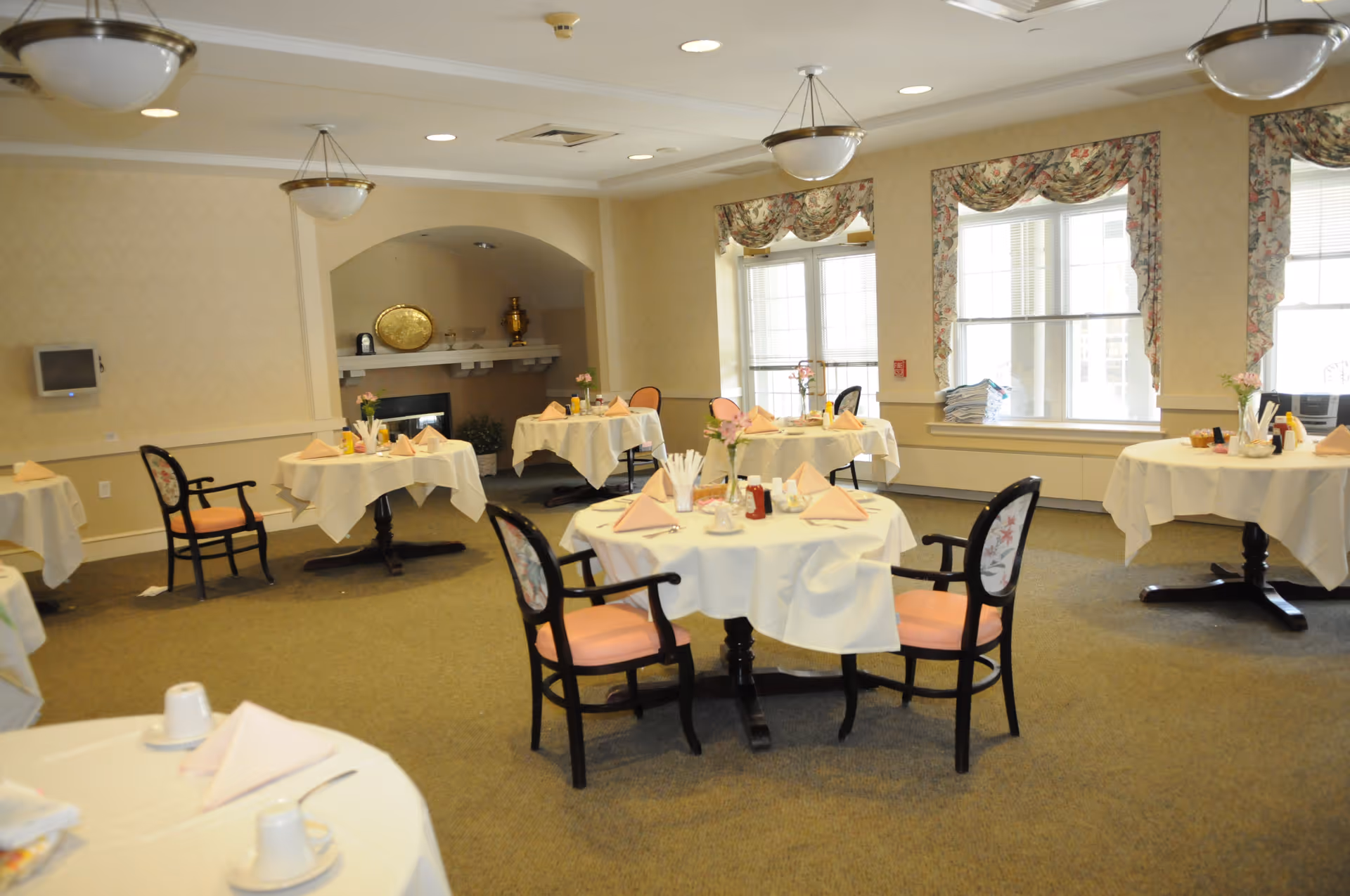 A dining room with round tables covered in white tablecloths, each set with folded napkins, cups, and small flower vases. The room has beige walls, carpeted floor, floral curtains on large windows, and ceiling light fixtures. There is a fireplace with decorative items on the mantel in the background.