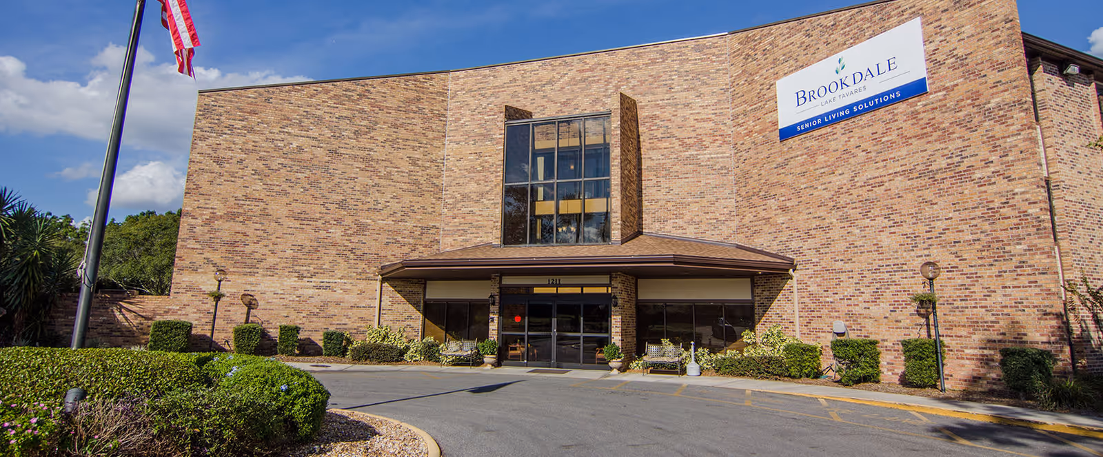 Exterior front view of Brookdale Lake Tavares senior living facility with a brick facade, large windows, an entrance with glass doors, landscaping, and an American flag on a flagpole.