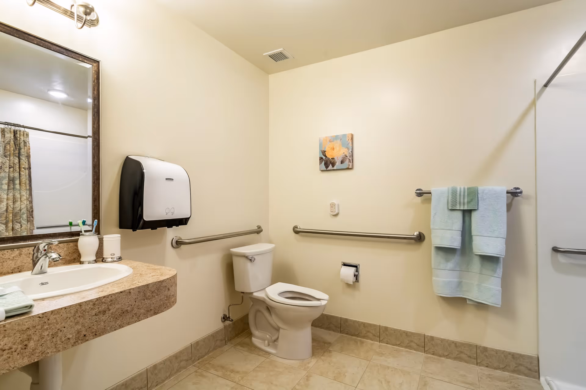 A clean and accessible bathroom with beige walls and tiled floor. The room features a toilet with grab bars on both sides, a wall-mounted paper towel dispenser, a sink with a granite countertop, a mirror above the sink, and a towel rack holding two folded towels. A small floral painting is hung on the wall above the toilet paper holder.