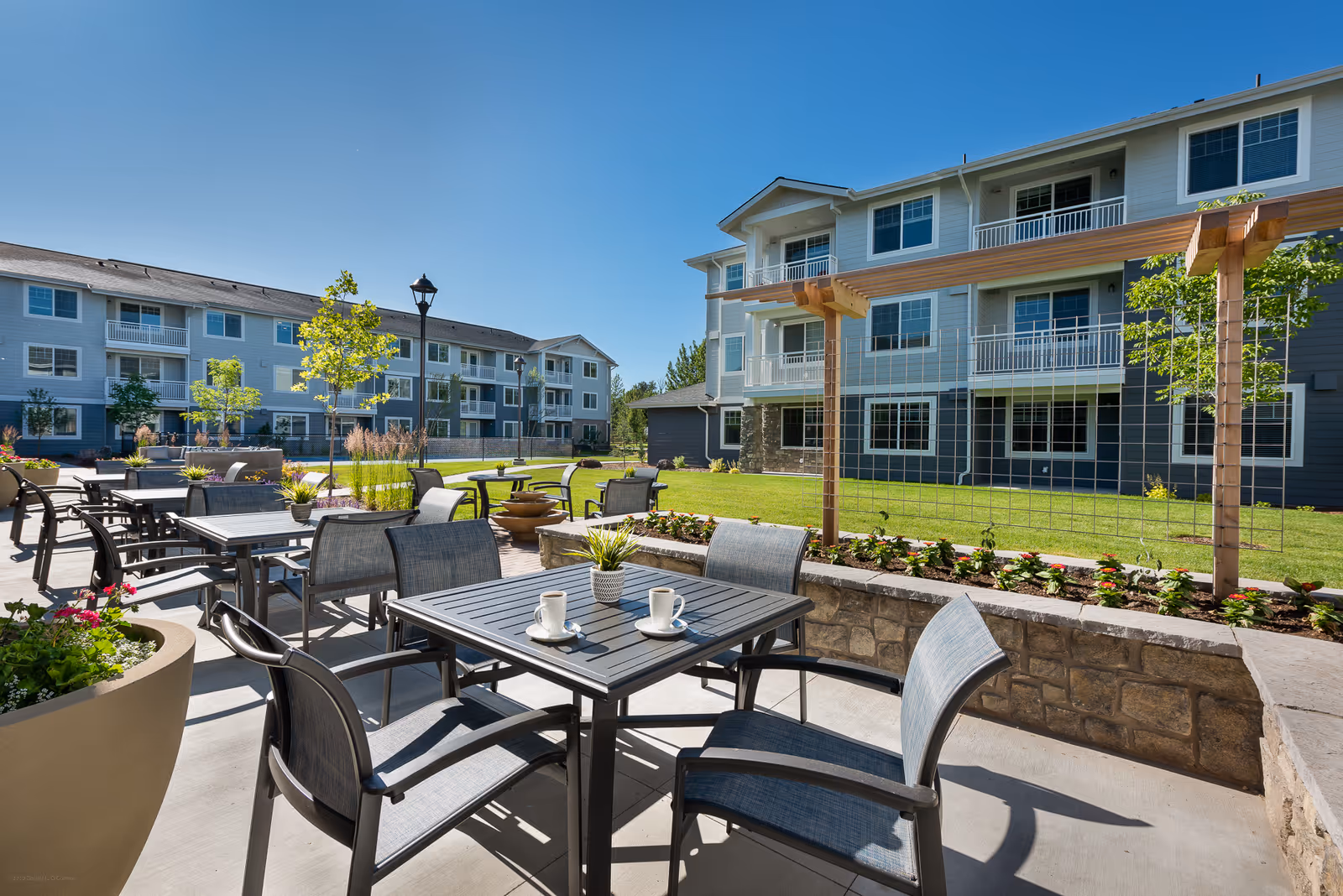 Outdoor patio area at Revel Eagle featuring multiple tables and chairs with potted plants and coffee cups on the tables, surrounded by well-maintained grass, young trees, and a trellis structure, with a three-story residential building in the background under a clear blue sky.