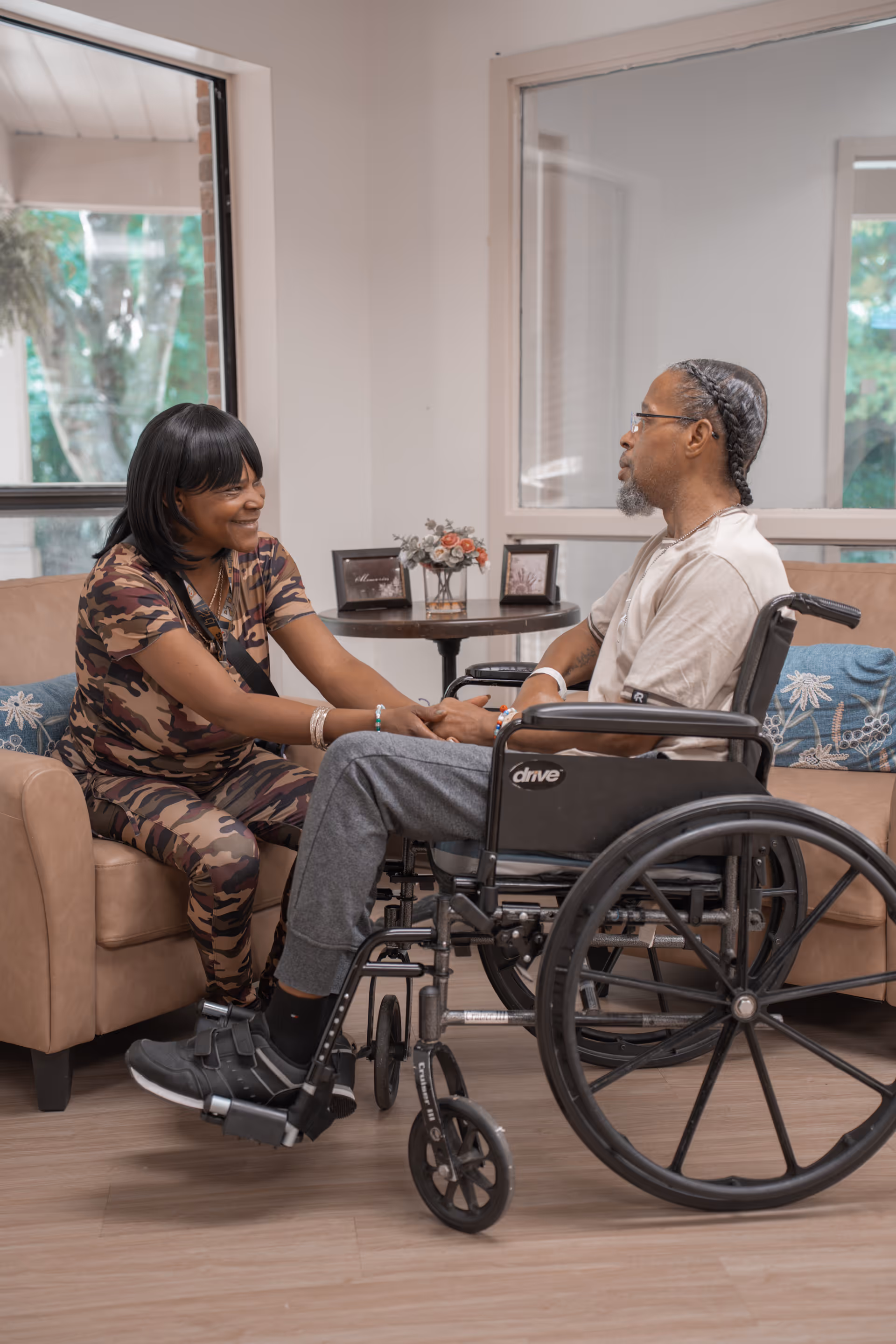 A woman sitting on a beige couch holding hands and smiling at a man in a wheelchair in a well-lit room with large windows and a small table with flowers and framed pictures in the background.