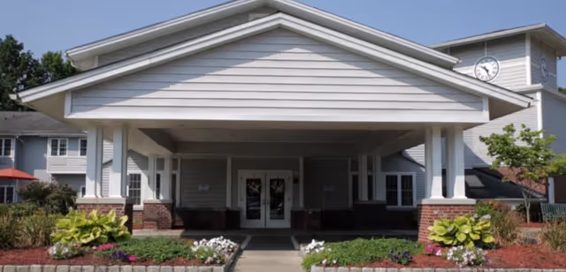Front exterior view of CareOne at Wayne Assisted Living facility showing a covered entrance with white pillars, brick accents, landscaped flower beds, and a clock tower on the right side of the building.