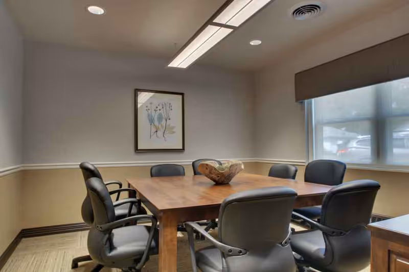 A conference room with a wooden table surrounded by eight black office chairs. The room has beige walls with a white chair rail, a framed floral artwork on the wall, a large window with a brown valance, and overhead fluorescent lighting.
