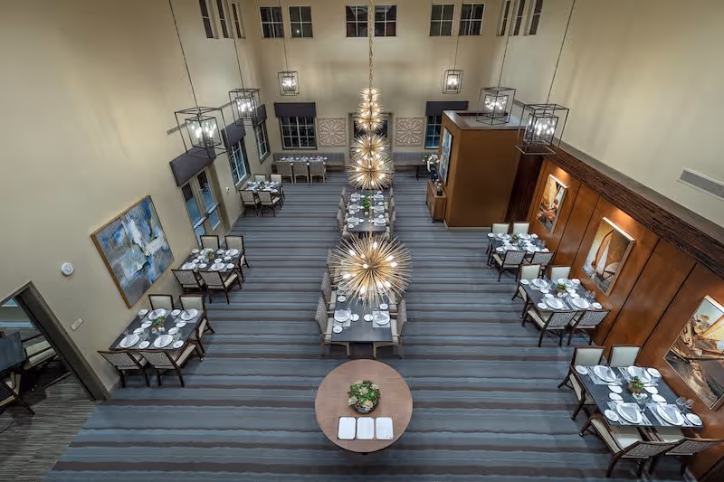 A spacious dining room in a senior living facility with multiple tables set for meals. The room features modern chandeliers, large windows, and artwork on the walls. The tables are arranged neatly with plates, glasses, and silverware, and there is a round table with a plant centerpiece in the foreground.