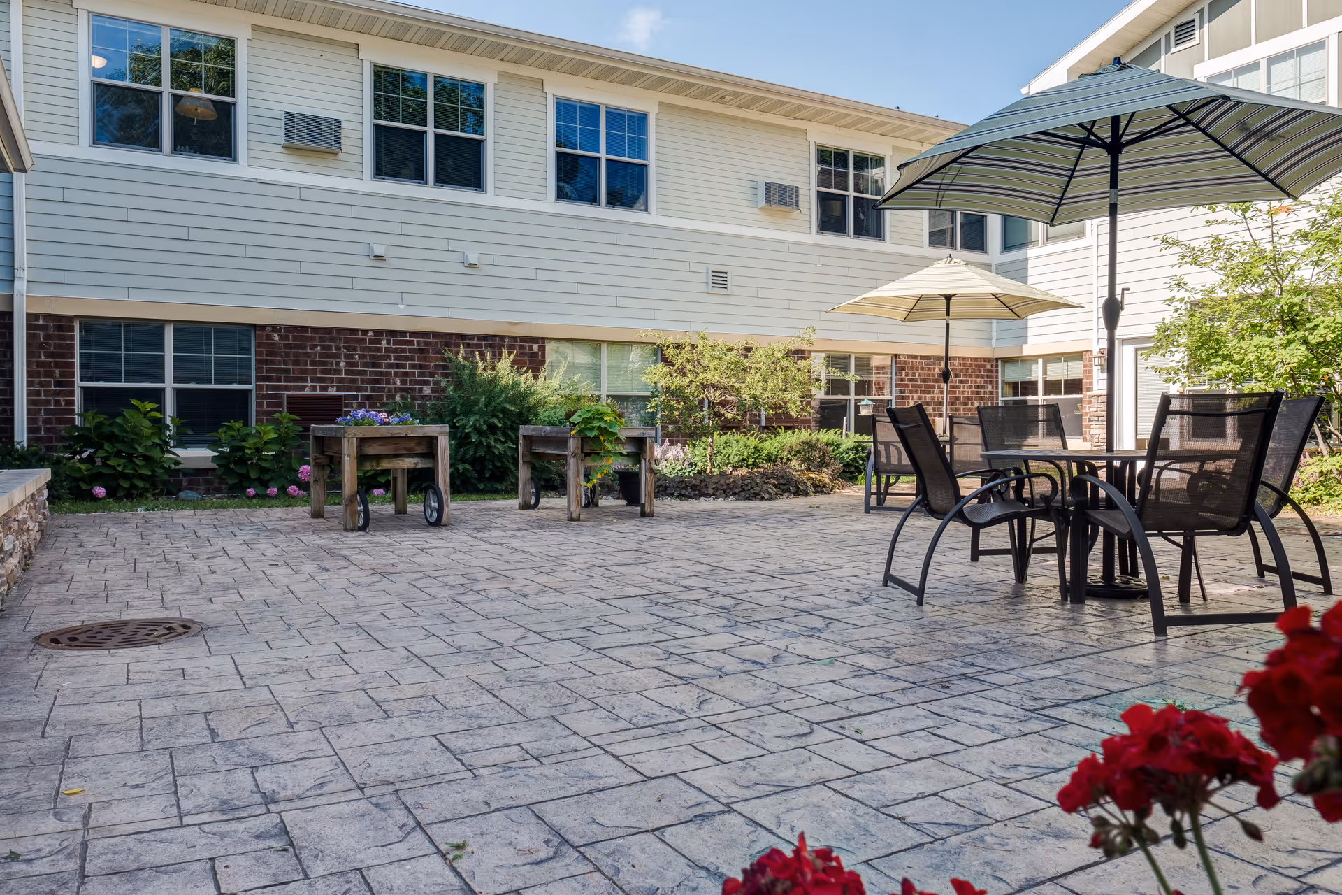 Outdoor patio area at Heritage Senior Campus with stone tile flooring, several tables with umbrellas, chairs, and raised garden beds. The building exterior features white siding and brick with multiple windows. Some greenery and flowers are visible around the patio.