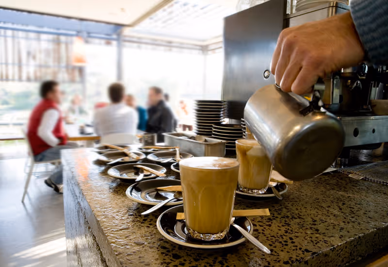 A barista pours milk into two lattes on a counter lined with saucers and spoons, with blurred patrons seated in a café in the background.