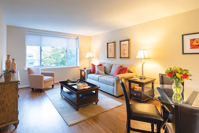 A bright, furnished living room with a sofa, armchair, coffee table, and dining table beside a large window.