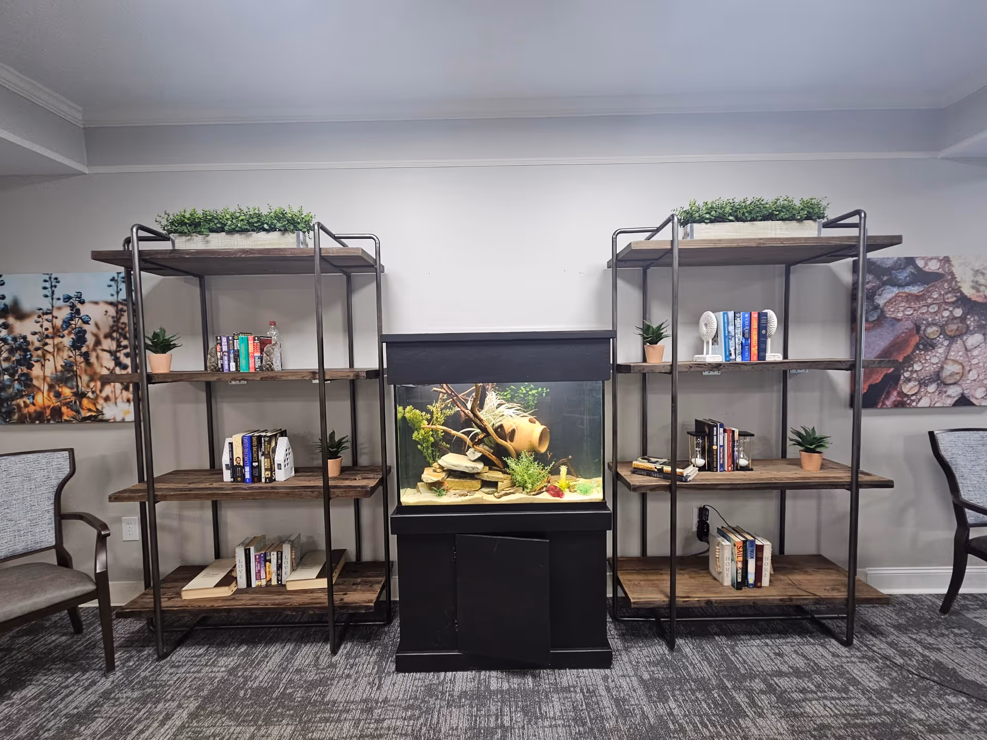 A room with two wooden and metal bookshelves on either side of a black fish tank filled with plants and decorative rocks. Each bookshelf has small potted plants and books arranged on the shelves. There are two chairs with wooden arms and gray cushions on each side of the bookshelves. The walls are light gray with two framed pictures featuring nature scenes.