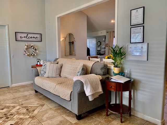 A cozy living room area with a gray couch covered by a beige quilted throw and several pillows. A white blanket is draped over one arm of the couch. Next to the couch is a small round wooden side table with a potted plant, a glass bottle, and some papers. On the wall above the table are three framed certificates or documents. To the left, a wall features a decorative wreath and a sign that says 'welcome to our home'. The floor is tiled with a beige and brown pattern. In the background, there is a glimpse of a dining area with chairs and a window with blinds.