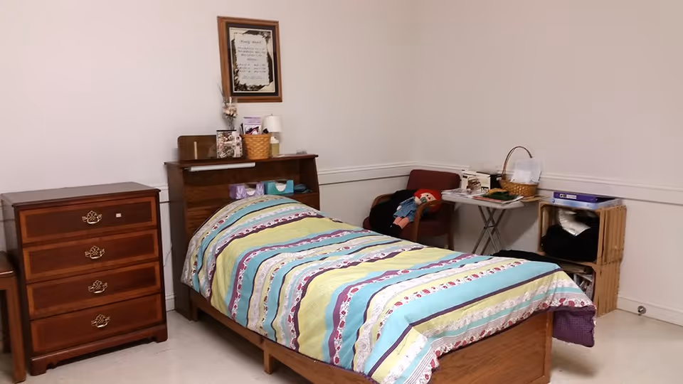 A simple senior bedroom featuring a bed, dresser, and a small table.