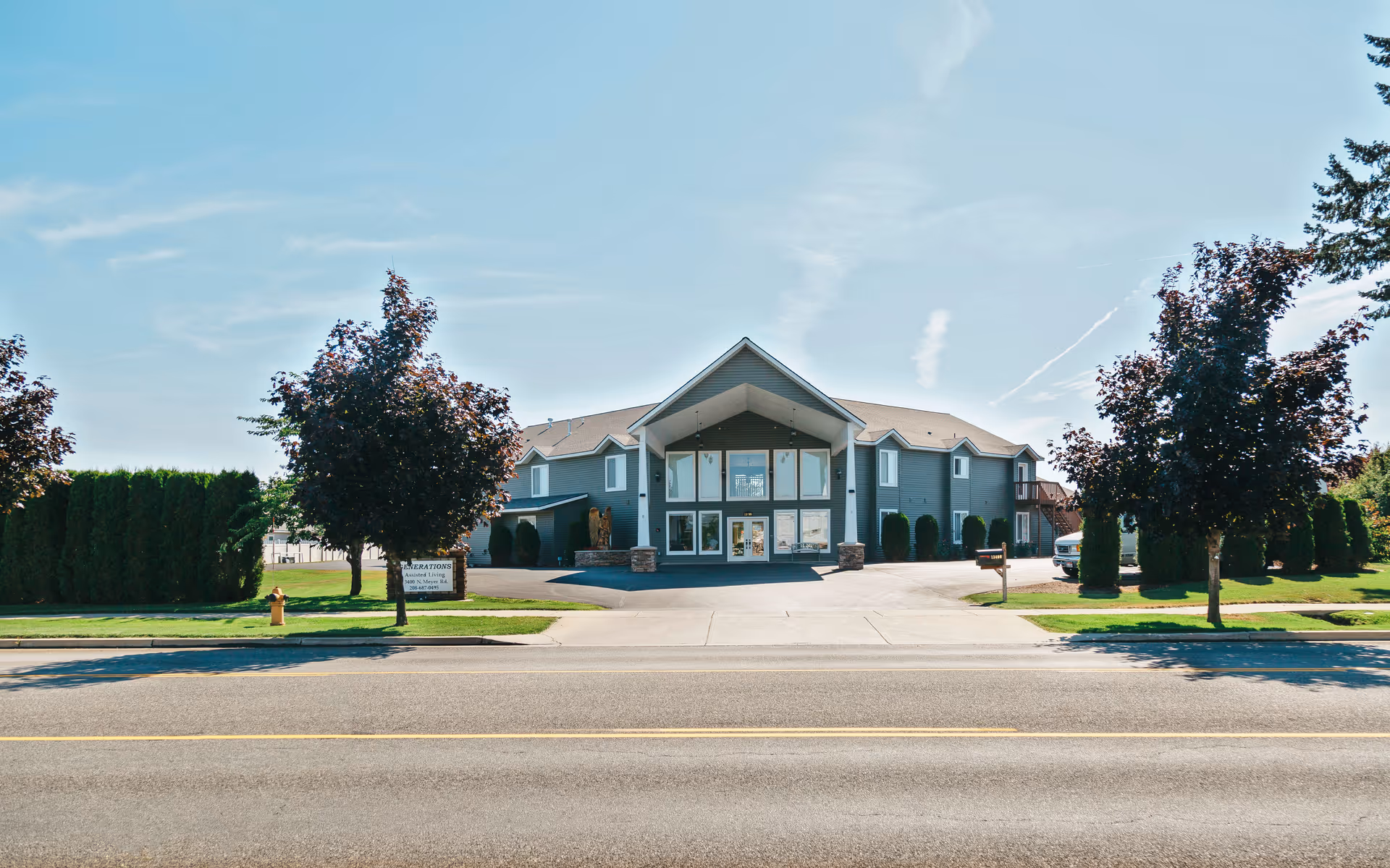 Front exterior view of a two-story building with a peaked roof and large windows at the entrance, surrounded by trees and greenery on a sunny day.