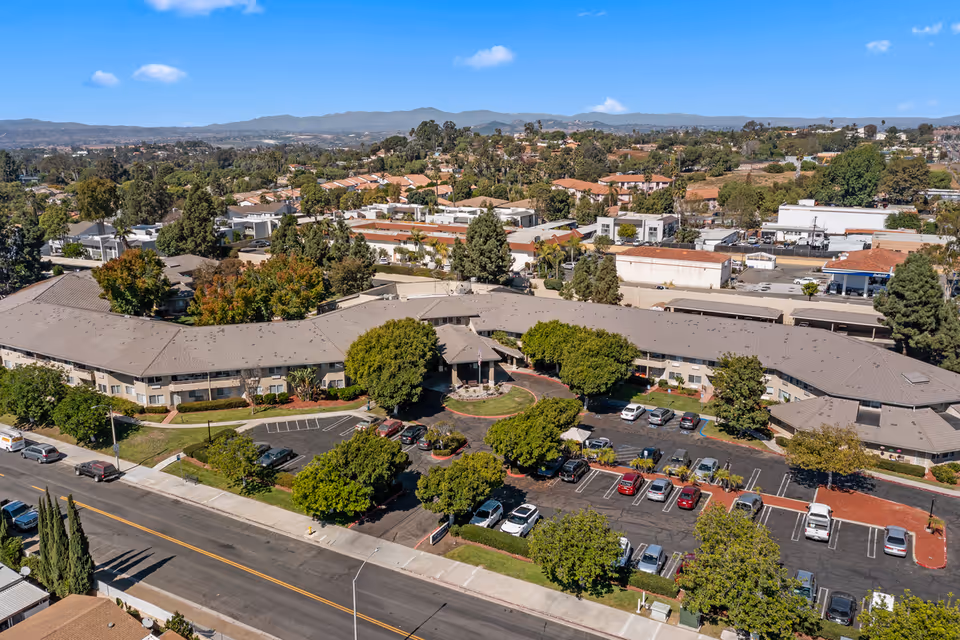 Aerial view of Arcadia Place Senior Living facility showing a large, single-story building with a gray roof surrounded by trees and parking lots. The facility is located in a suburban area with other buildings and hills visible in the background under a clear blue sky.