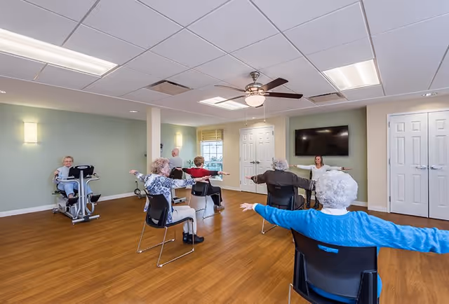 A group of elderly individuals seated on chairs in a spacious room with wooden floors and light green walls, participating in a seated exercise class led by an instructor at the front near a wall-mounted TV. One person is using an exercise machine on the left side of the room.
