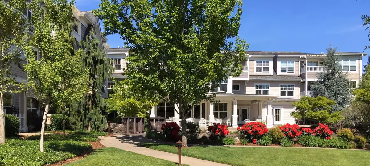 Exterior view of The Stafford Retirement Community building with a well-maintained garden featuring green trees, red flowering bushes, and a paved walkway under a clear blue sky.