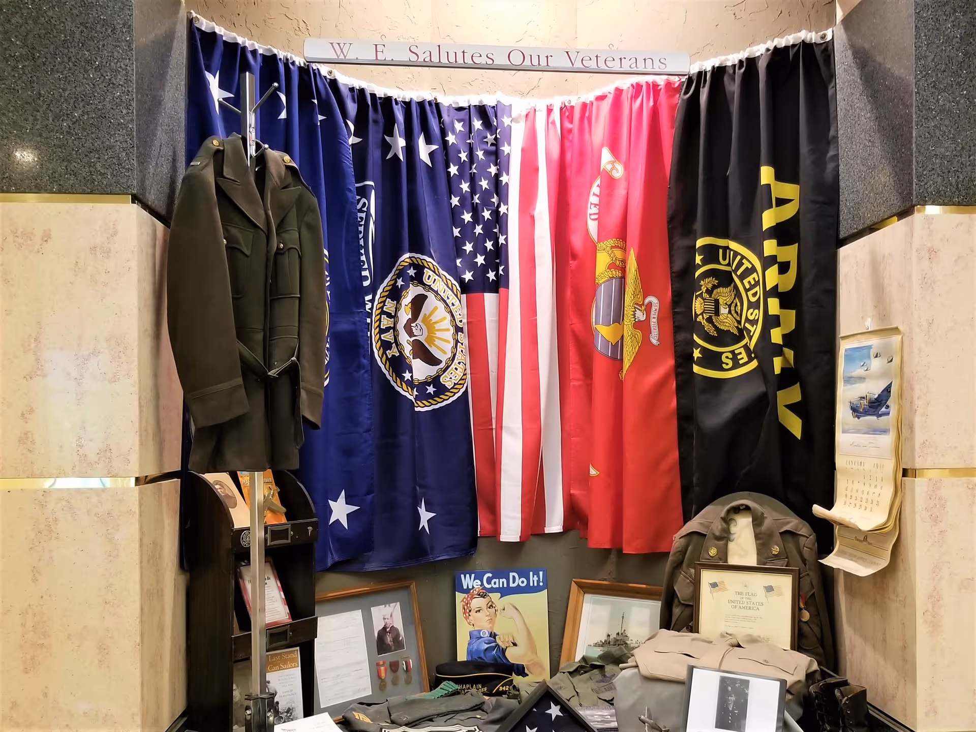 A veterans tribute display featuring military flags, a coat rack with a military jacket, framed photographs, medals, a 'We Can Do It!' poster, and various military memorabilia arranged in a corner with a sign above reading 'W. E. Salutes Our Veterans'.