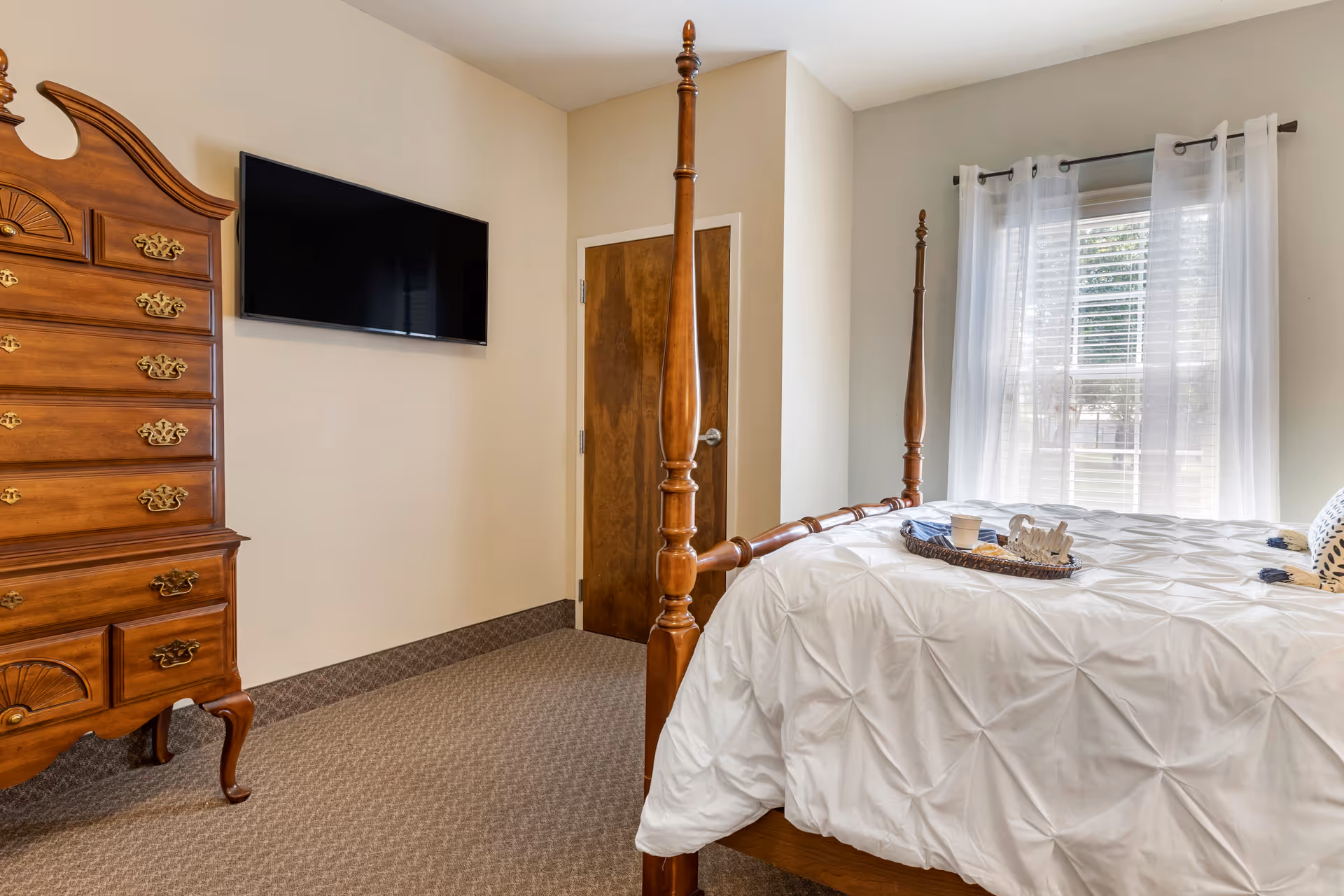 A bedroom in Wesley Court Assisted Living Community featuring a wooden four-poster bed with white bedding, a tray with a cup and decorative items on the bed, a wooden dresser with brass handles, a wall-mounted flat-screen TV, a wooden door, and a window with sheer white curtains letting in natural light.