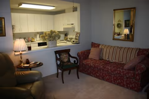 Small living room with a patterned red sofa, leather recliner, side table and lamp, and a kitchen visible through a pass-through window.
