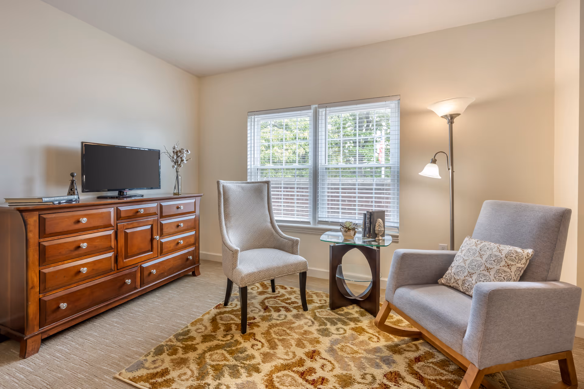 Well-lit sitting area with two armchairs, a wooden dresser with a TV, a side table and floor lamp in front of a window.