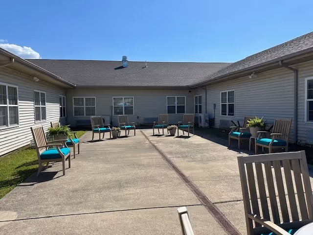 Sunny enclosed courtyard with wooden chairs featuring teal cushions arranged around a central concrete patio between single-story building wings.