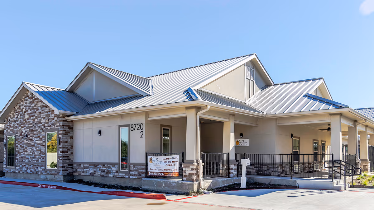 Exterior view of a single-story senior living facility building with a metal roof, beige and brick facade, and a covered entrance with railings. A sign near the entrance reads 'No More Wait!! We are now Open!!! Assisted Living Memory Care' along with a phone number. The building number 8720 2 is visible on the wall.