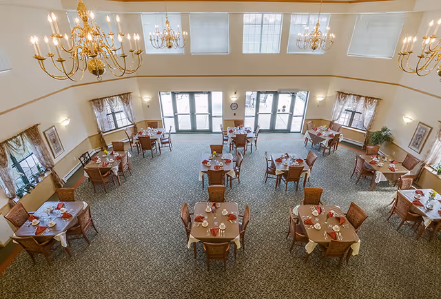 A spacious dining room with multiple square tables set for meals, each with chairs, napkins, and tableware. The room has large windows and glass doors letting in natural light, elegant chandeliers hanging from the ceiling, and patterned carpet flooring.