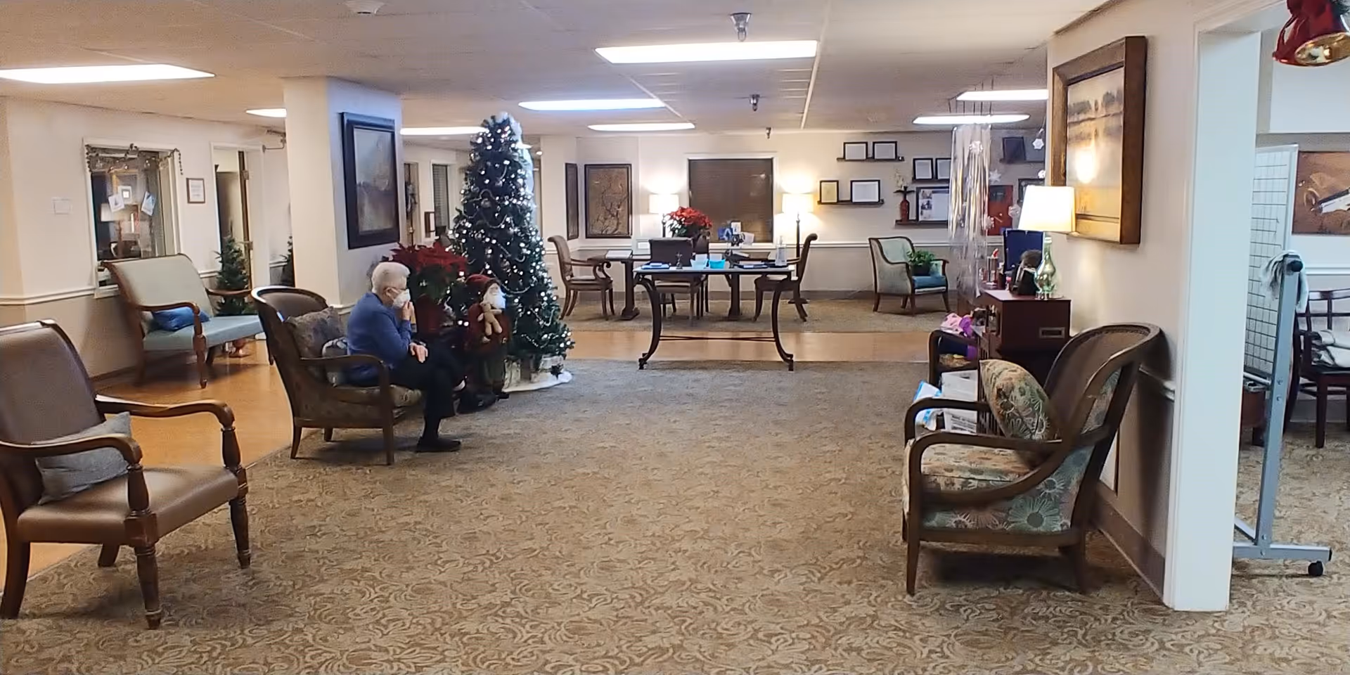 A senior living facility common area decorated for the holidays with a Christmas tree and poinsettias. Several chairs are arranged around the room, and an elderly person is sitting on one of the chairs. The room has a carpeted floor and is well-lit with ceiling lights and lamps. There are tables and framed pictures on the walls in the background.
