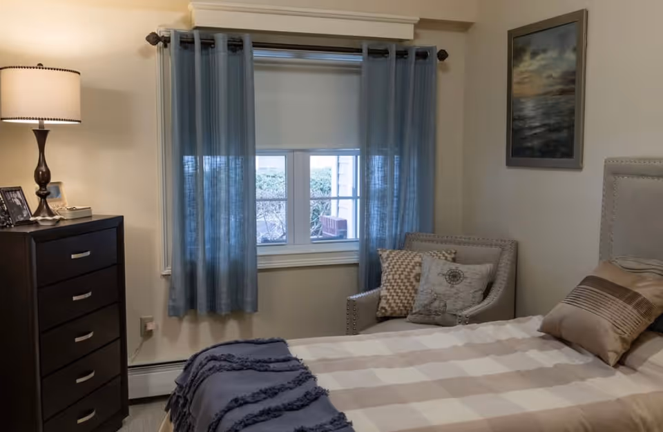 A cozy bedroom with a bed covered in a beige and white checkered blanket and several pillows. Next to the bed is a cushioned armchair with decorative pillows. A window with blue curtains and a white roller shade is centered on the wall. To the left, there is a dark wooden dresser with a lamp and framed photos on top. A framed painting hangs on the wall to the right.