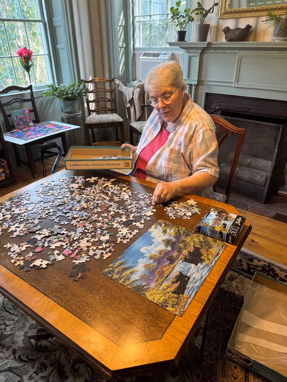 An elderly woman sitting at a wooden table working on a jigsaw puzzle in a cozy room with large windows, plants, and a fireplace. The puzzle box and partially completed puzzle are on the table, and the woman is holding a puzzle piece.