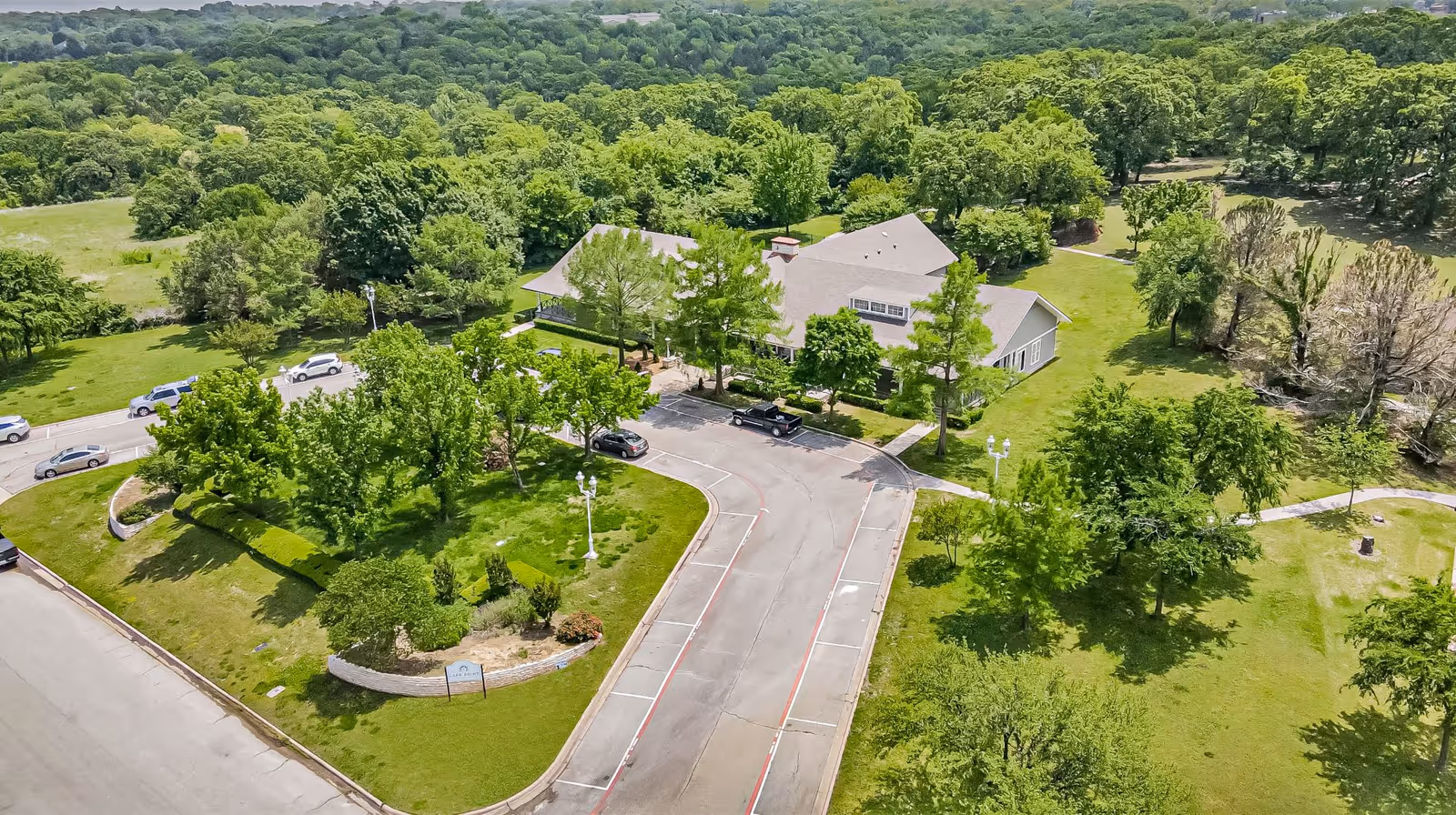 Aerial view of The Homestead of Denison facility surrounded by lush green trees and landscaping. The building has a light-colored roof and is situated in a spacious area with a driveway and parking spaces. The surrounding area is filled with dense greenery and open grassy spaces.
