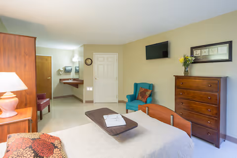 Well-lit senior care bedroom with a bed and overbed table, wooden dresser, wall-mounted TV, and a teal armchair.