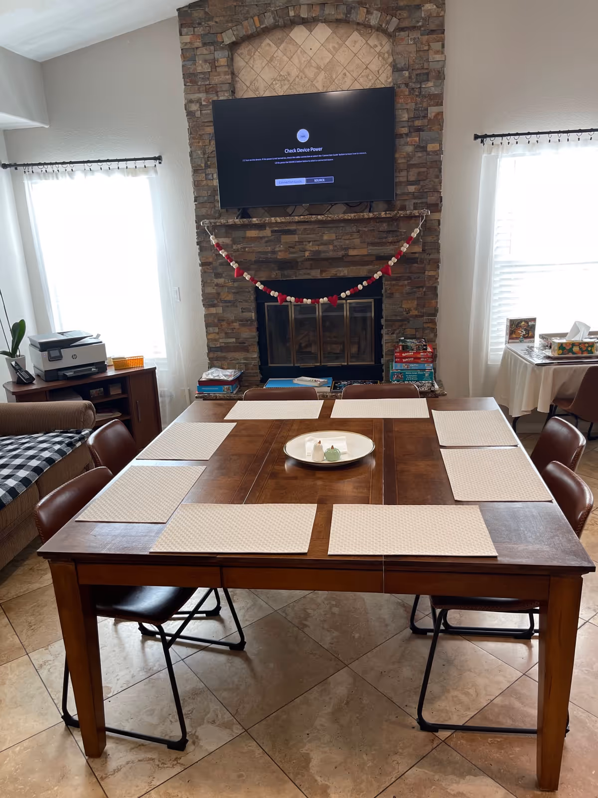 Wood dining table set with placemats in a living area facing a stone fireplace with a mounted TV.