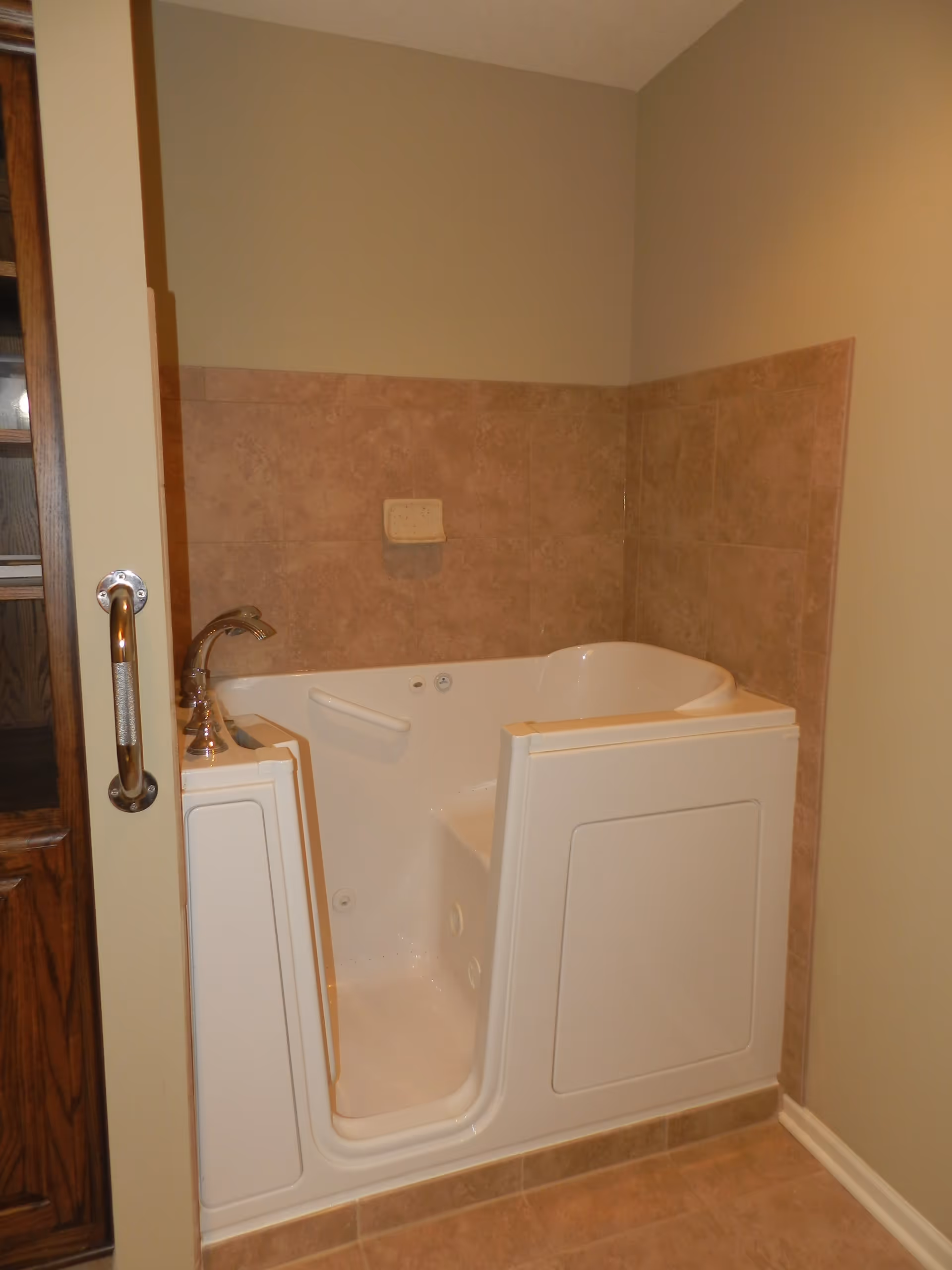 A walk-in bathtub with its door open, faucet, grab bar, and tiled surround in a bathroom.