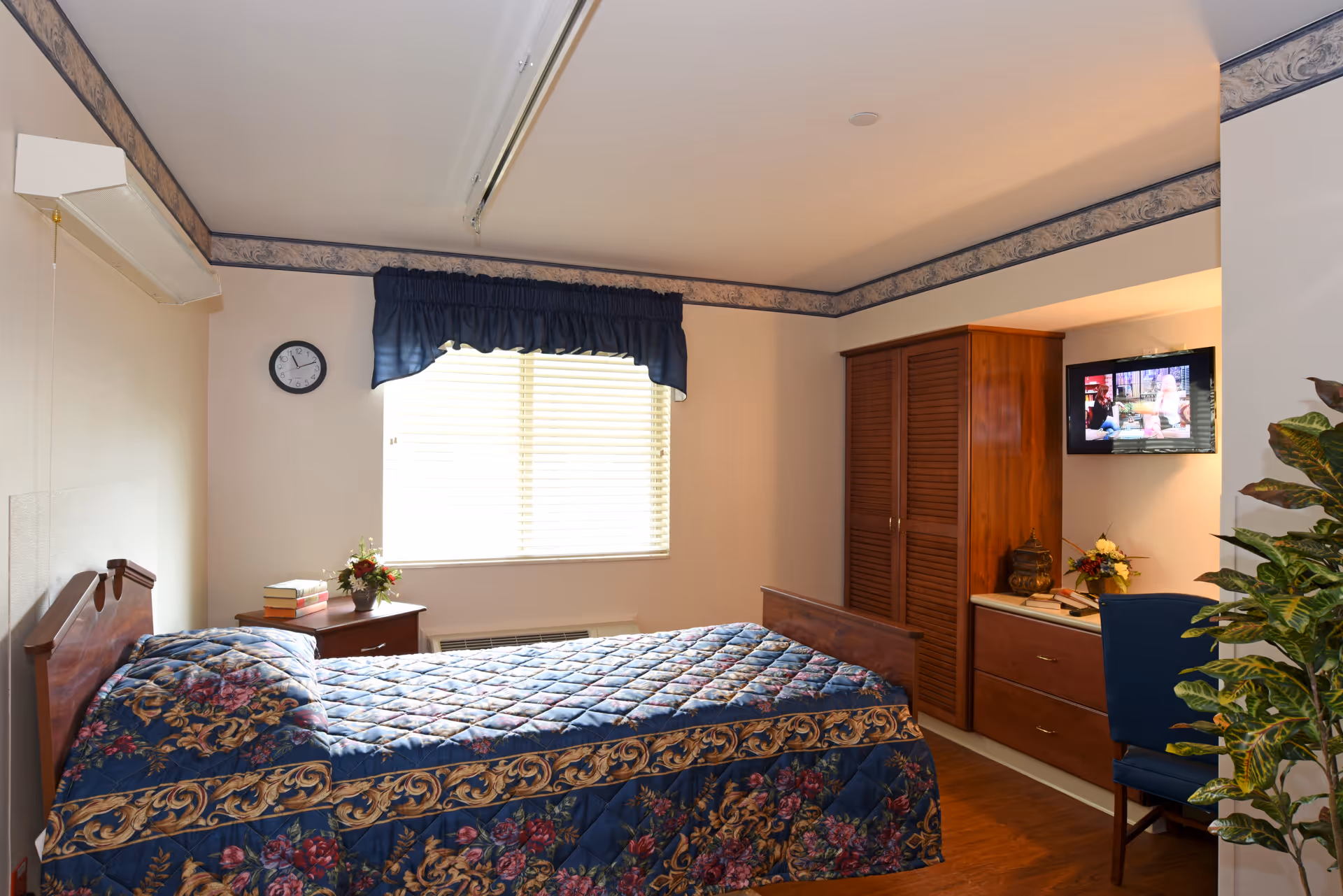 A cozy bedroom in a senior living facility with a single bed covered in a blue floral quilt. There is a wooden nightstand with books and a flower vase next to the bed. A window with blinds and a dark blue valance lets in natural light. On the opposite wall, there is a wooden wardrobe and a small dresser with a chair and a mounted TV above it. A plant is partially visible on the right side of the image.