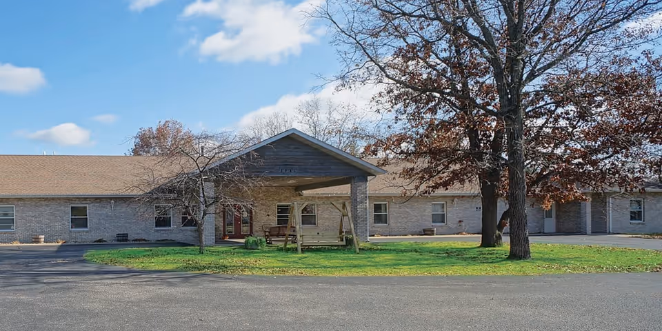 Single-story brick senior living building with a covered entrance, a wooden swing bench, trees, and a paved driveway.