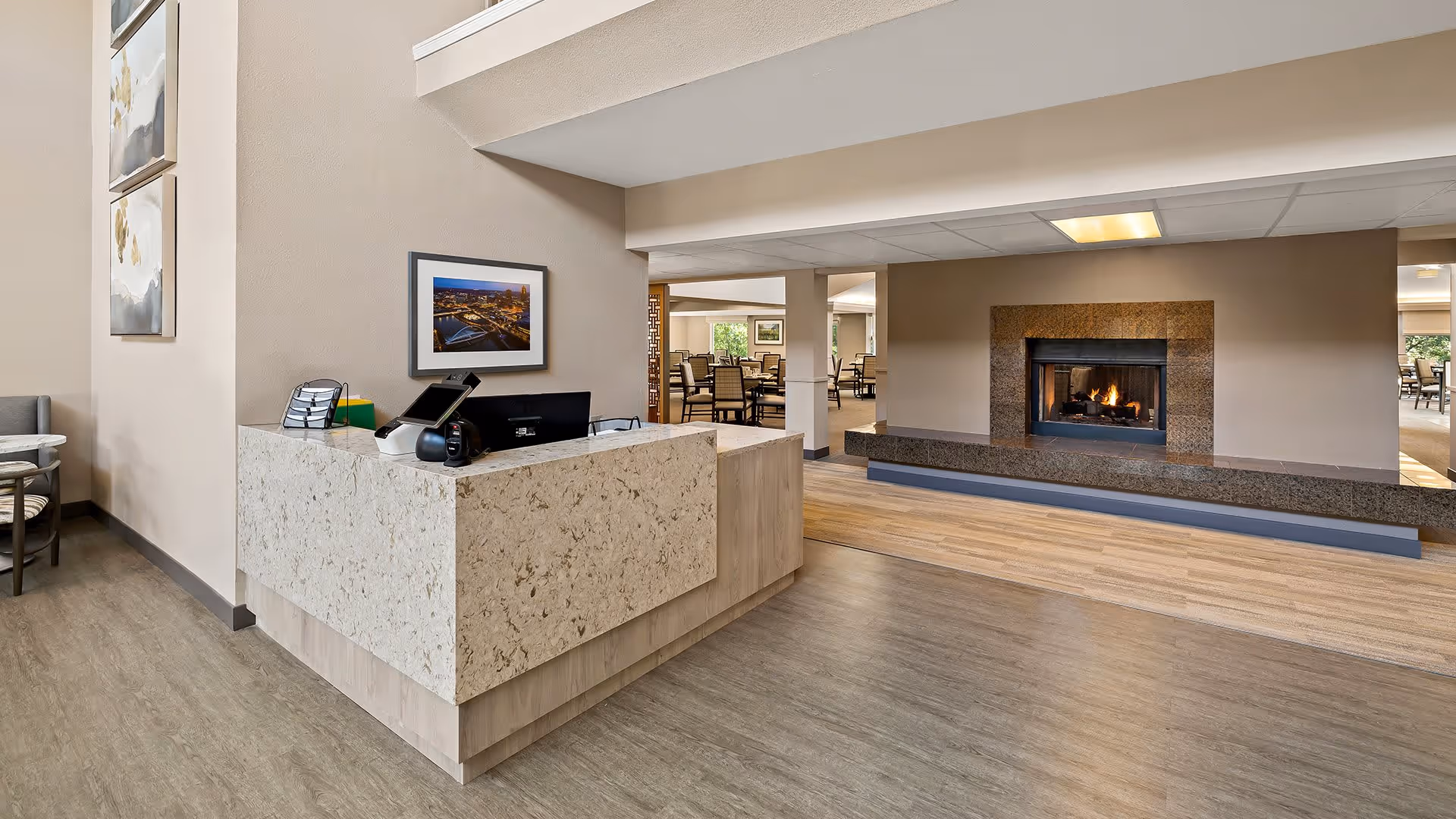 Reception desk area with a granite countertop in a senior living facility. Behind the desk is a framed picture on the wall. To the right, there is a cozy fireplace with a fire burning, surrounded by a granite hearth. In the background, there is a dining area with tables and chairs visible through open doorways. The flooring transitions from a wood-like material near the fireplace to a grayish floor near the reception desk.