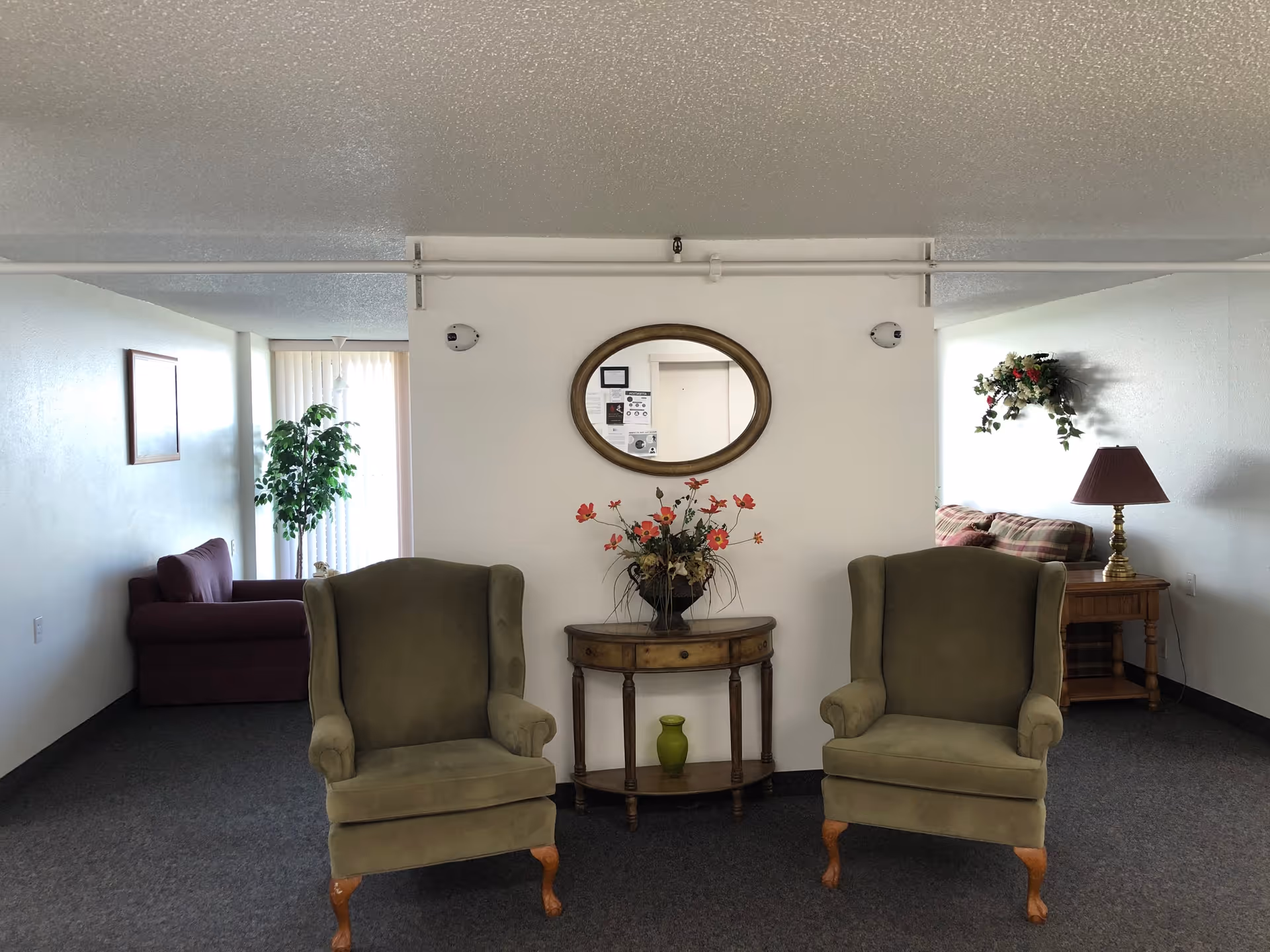 A sitting area with two green upholstered armchairs flanking a small wooden table topped with flowers beneath an oval mirror in a senior living common room.