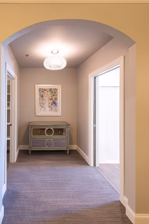 A hallway with beige walls and carpeted floor leading to a small decorative chest of drawers with circular designs. Above the chest hangs a framed abstract artwork. There is a ceiling light fixture with a crystal-like design. Doorways on either side of the hallway lead to other rooms, one with a white curtain visible.