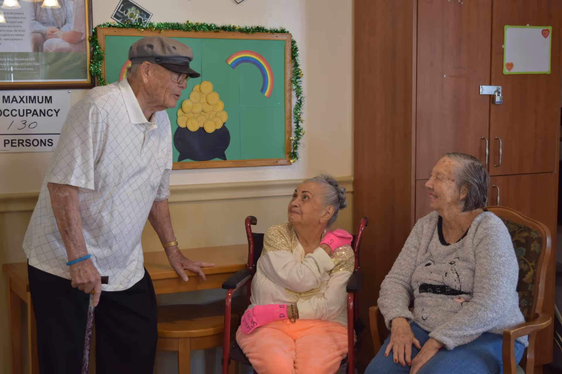 Three elderly individuals in a room at Rose Garden Residential Care. One man wearing a cap and glasses is standing and leaning on a cane, talking to two seated women. One woman is in a wheelchair wearing bright pink gloves and looking up at the man, while the other woman in a gray sweater is seated in a chair smiling. Behind them is a green bulletin board decorated with a rainbow and a pot of gold, and wooden cabinets.