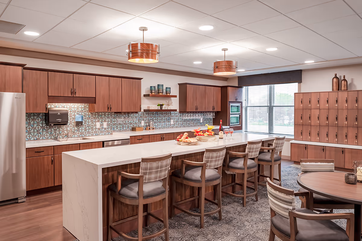 A modern kitchen area with wooden cabinets, a large white marble island with five cushioned bar stools, and a tiled backsplash. The kitchen features stainless steel appliances, including a refrigerator and built-in ovens. There are decorative items on the shelves and countertop, and a window with blinds lets in natural light. A round table with chairs is visible in the foreground.
