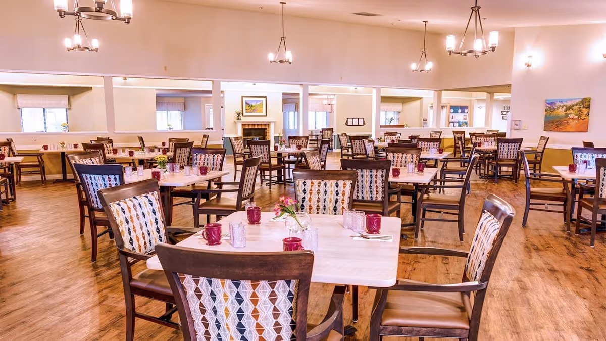 A spacious dining room with multiple square tables and wooden chairs featuring patterned upholstery. Each table is set with purple mugs, glasses, and small flower arrangements. The room has wooden flooring, light-colored walls, several hanging light fixtures, and large windows with blinds allowing natural light to enter.