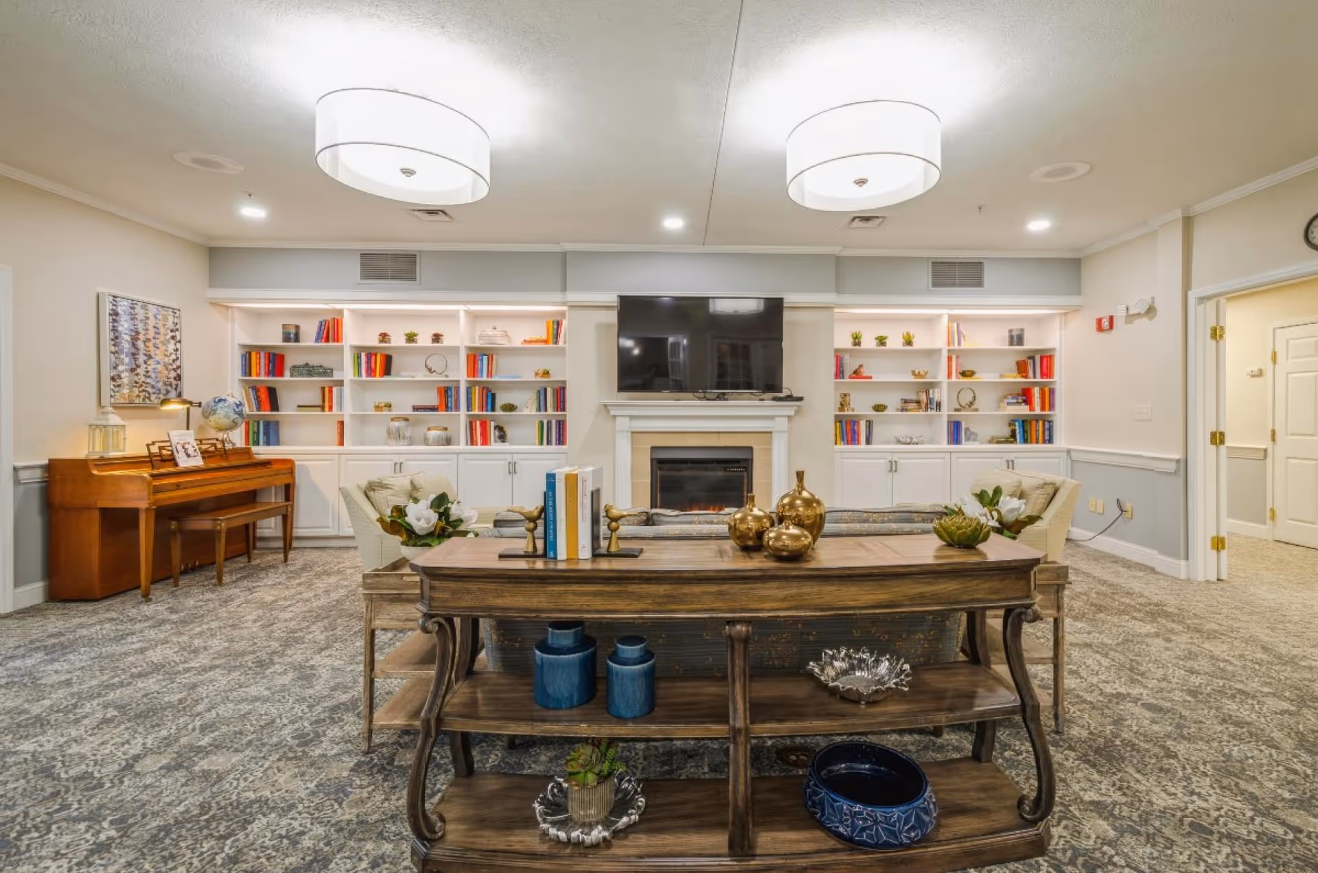 A furnished communal living room with built-in bookshelves, a fireplace and television, piano and seating.